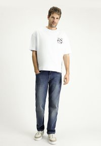 Camiseta de algodón blanca con un gráfico en el lado izquierdo del pecho, combinada con jeans de mezclilla azules. El modelo lleva zapatillas blancas y grises.