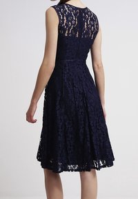 Navy blue lace dress with sleeveless design, floral pattern, flared skirt, and sheer upper back. Textured fabric with a fitted waist.
