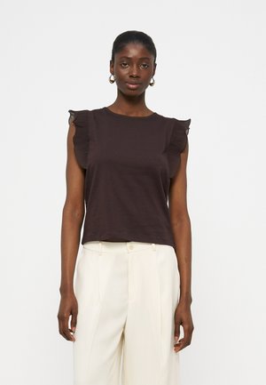 Γυναίκα με μαύρο αμάνικο τοπ με βολάν στους ώμους και λευκό ψηλόμεσο παντελόνι, στέκεται μπροστά σε ένα απλό φωτεινό φόντο.