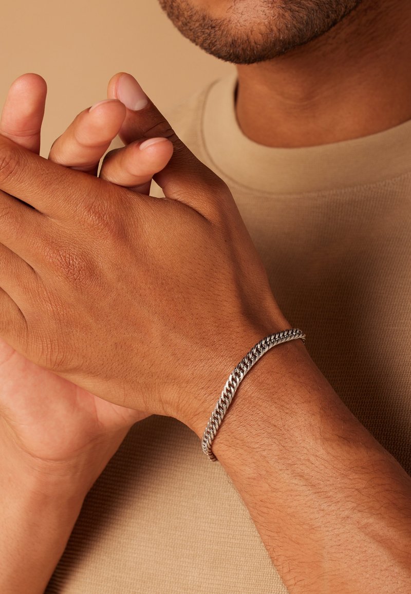 Zilveren schakelarmband met een gedraaid ontwerp, liggend op een bruine huidtoon pols, tegen een gestructureerde beige achtergrond.