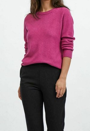 Vrouw draagt een nauwsluitende magenta trui met geribbelde manchetten en zwarte broek met hoge taille, tegen een effen lichte achtergrond.