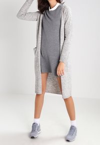 Lungo cardigan grigio con texture a coste, apertura frontale e due tasche laterali, indossato sopra un vestito grigio attillato e scarpe da ginnastica grigie.