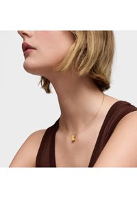 Collana d'oro con un piccolo pendente a forma di orsetto su una catena sottile, indossata sopra un top marrone, mettendo in risalto il suo design delicato.