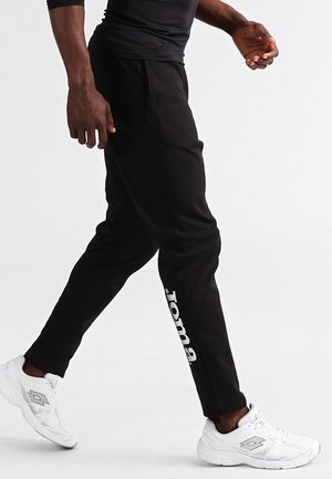 Mann läuft, trägt schwarze Jogginghosen mit weißem Logotext am Bein und weiße Sportschuhe vor weißem Hintergrund.