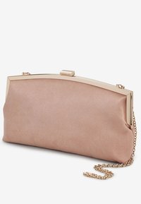 Pochette en suède rose pâle avec cadre en métal doré et bandoulière chaîne, reposant sur une surface blanche en léger angle.