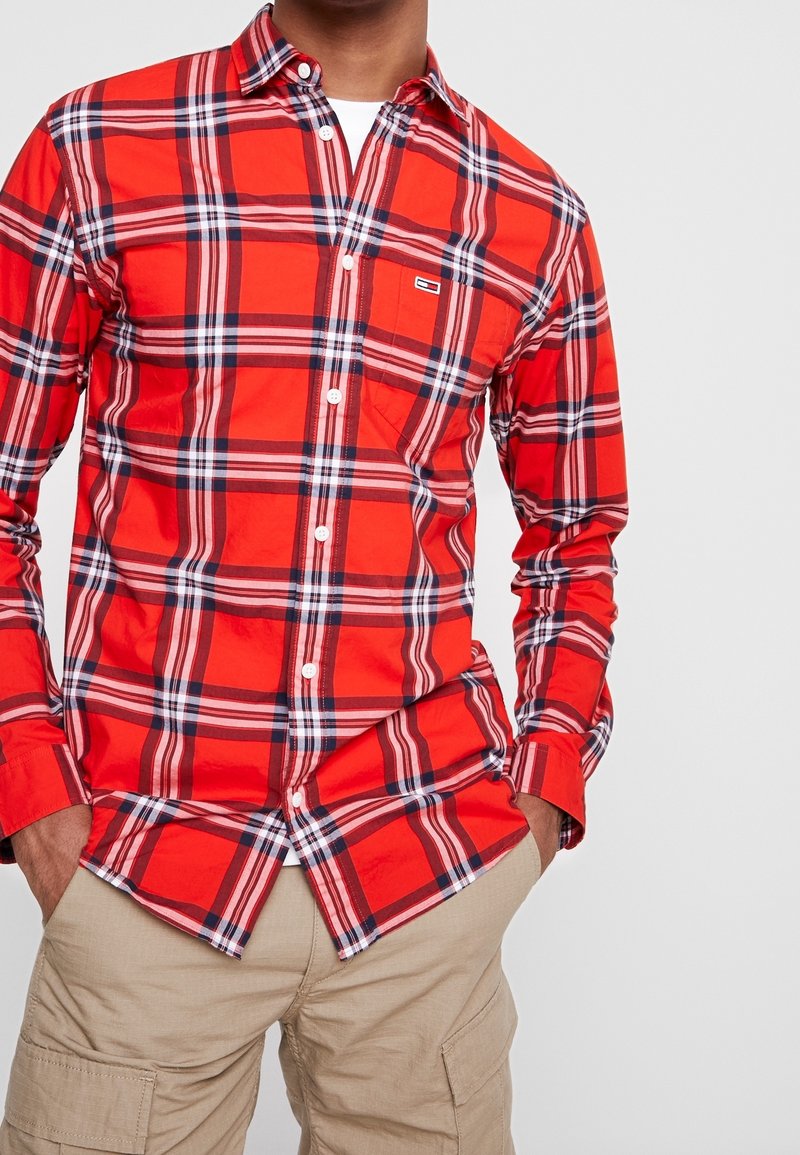 Chemise à carreaux rouges à manches longues, boutons blancs et poche poitrine ; présente des motifs à carreaux bleus et blancs ; associée à un short kaki.