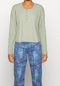 Ljusgrön långärmad henley-topp med fyra knappar, matchad med blå mönstrade vida byxor som har en collage av ögonmönster.