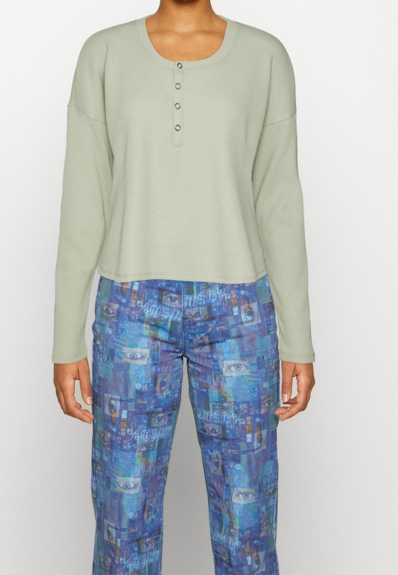 Ljusgrön långärmad henley-topp med fyra knappar, matchad med blå mönstrade vida byxor som har en collage av ögonmönster.