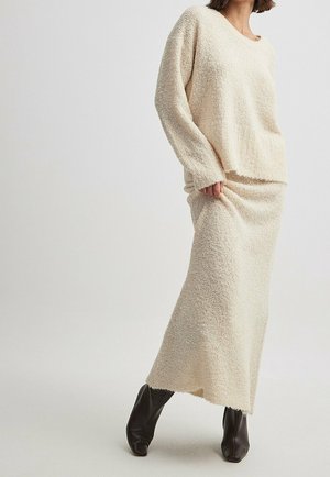 Femme portant un pull en maille crème et une longue jupe assortie, associée à des bottines noires, debout devant un fond clair uni.