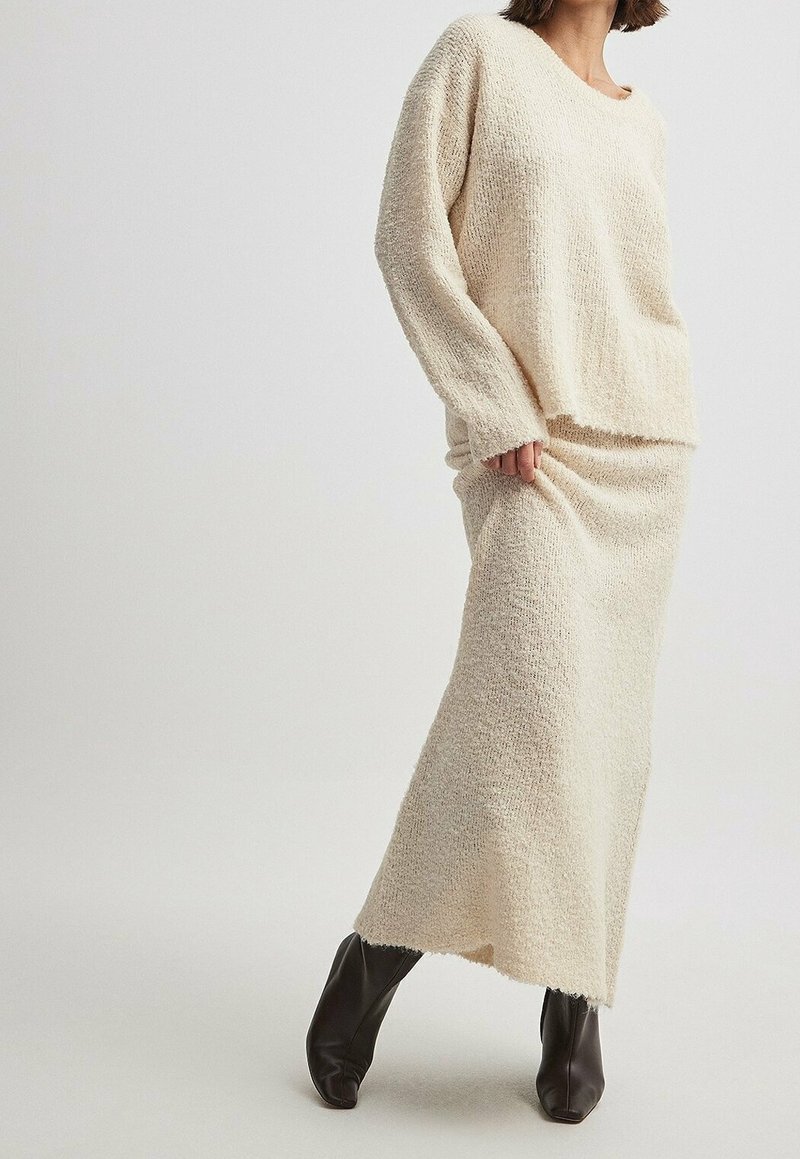 Femme portant un pull en maille crème et une longue jupe assortie, associée à des bottines noires, debout devant un fond clair uni.