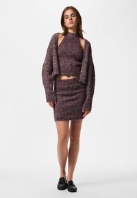 Jeune femme portant un crop top en maille violette, un cardigan et une jupe assortis, avec des mocassins noirs, debout devant un fond blanc uni.