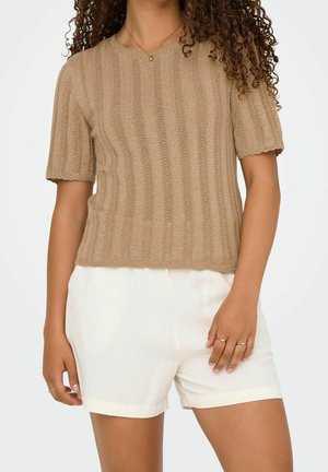 Vrouw draagt een beige gebreide top met korte mouwen en witte shorts met hoge taille, met krullend haar en eenvoudige gouden sieraden.