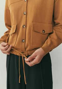 Cardigan maro tricotat, cu nasturi în față, două buzunare la piept și tiv cu șnur, purtat peste pantaloni negri. Materialul pare moale și texturat.