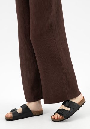 Füße in schwarzen Doppelriemen-Slides mit Korksohlen und braunen, strukturierten, weit geschnittenen Hosen vor einem einfarbigen Hintergrund.