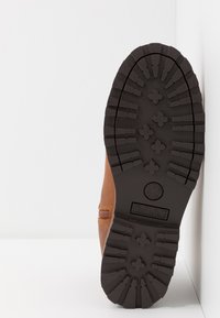 Botte en cuir marron avec une semelle en caoutchouc noir robuste. Présente des rainures profondes et un logo subtilement embossé sur le côté. Construction solide.