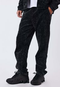 Persona che indossa pantaloni in corduroy neri, sneakers nere e una giacca nera abbinata, con una mano in tasca davanti a uno sfondo semplice.