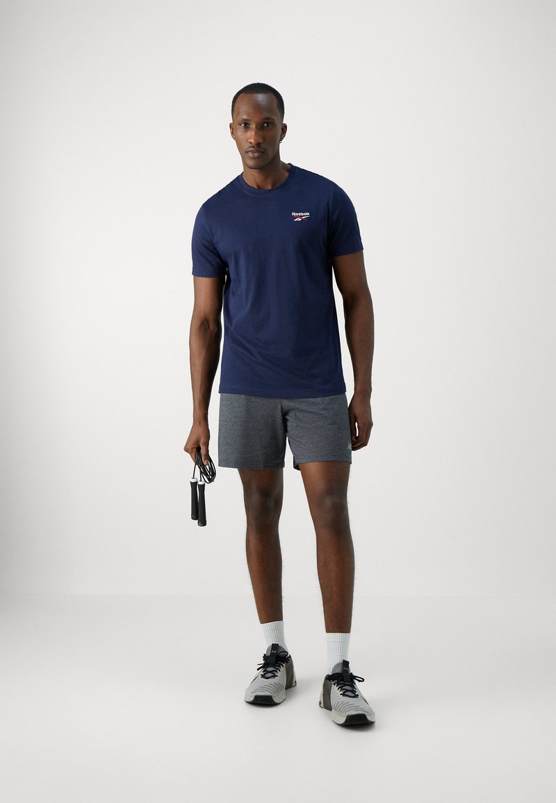 T-shirt de sport bleu marine à manches courtes avec un petit logo, associé à un short gris. Le modèle tient une corde à sauter et porte des baskets grises.