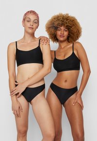 Two models wear matching black, seamless bralettes and bikini bottoms. The fabric appears smooth and stretchy, with adjustable straps.