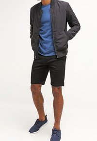Veste bomber noire sur un t-shirt bleu, associée à un short noir et des baskets bleu marine. Tissu lisse, coupe décontractée et design simple.