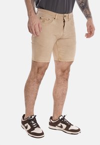 Shorts in denim beige con una forma aderente, cinque tasche e dettagli di cucitura. Indossati con sneakers marroni e bianche, che mostrano un look casual.