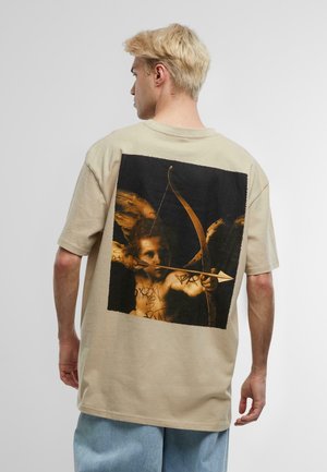 Beżowy t-shirt z dużą, ciemną grafiką aniołka z łukiem i strzałą z tyłu. Krótkie rękawy, luźny krój, miękki materiał bawełniany.