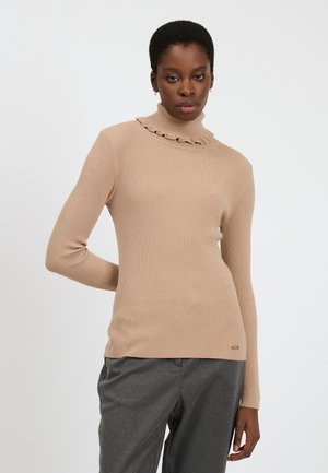 Vrouw in een beige geribbelde turtleneck trui met ruches en grijze broek, staand voor een effen witte achtergrond.