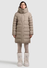 Béžová prešívaná parka s vysokým golierom, prednými gombíkmi a vreckom na zips; v kombinácii s bielymi snehovými topánkami a svetlými legínami.