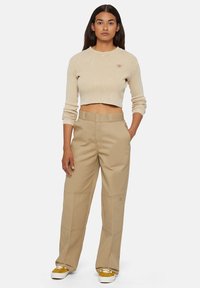 Beige cropped gebreide trui met lange mouwen, gecombineerd met beige wijde broek met naden en een logopatch. Sneakers met gele accenten.