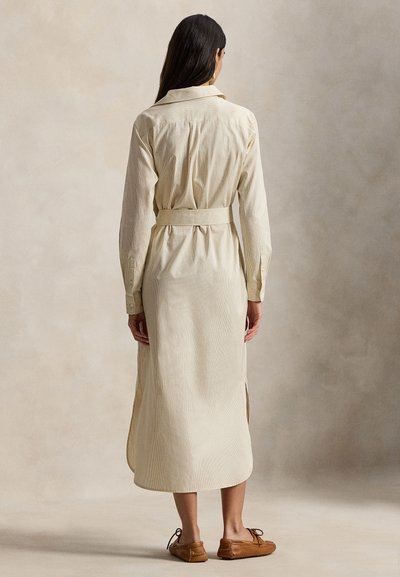 Μακρύ κρεμ πουκάμισο-φόρεμα με κάθετες ρίγες, ζώνη στη μέση, μυτερός γιακάς, μανσέτες με κουμπιά και πλευρικές σχισμές, συνδυασμένο με καφέ παπούτσια χωρίς κορδόνια.