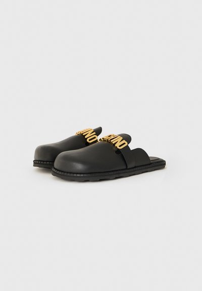 Svarta slip-on sandaler med tjocka sulor och gulddekorativa bokstäver över remmarna, mot en enkel vit bakgrund.