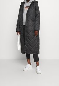 Persona che indossa un lungo cappotto trapuntato nero, una felpa grigia con cappuccio, jeans neri a vita alta, sneakers bianche e tiene in mano una piccola borsa bianca, in piedi su un pavimento grigio.