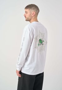 Weißes Langarm-T-Shirt mit einem Spinnennetzgrafik und einer grünen Figur auf dem Rücken, mit Textdetails auf den Ärmeln.