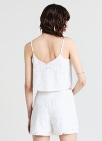 Weißes Zweiteiler-Outfit mit einem verkürzten Oberteil und Shorts. Der Stoff ist strukturiert und mit gestickten Mustern sowie Lochdetails verziert. Die Träger sind dünn.