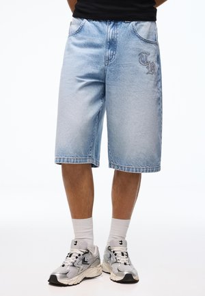 Mężczyzna noszący jasnoniebieskie dżinsowe szorty z haftem "TR", białe skarpetki i srebrno-szare sportowe buty, stojący na białym tle.