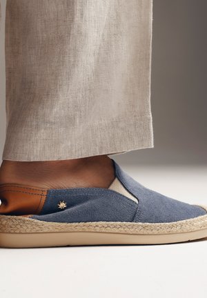 Piede che indossa pantaloni di lino beige e scarpa blu slip-on con tallone in pelle marrone chiaro e suola intrecciata in iuta, in piedi su uno sfondo neutro.