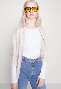 Leichte Leinenbluse über einem weißen T-Shirt, kombiniert mit hoch taillierten blauen Jeans. Große schwarze Sonnenbrillen mit gelben Gläsern runden den Look ab.