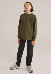 En ung person som står upp, klädd i en olivgrön långärmad skjorta med texten "BLUE RIDGE", svarta jeans och beige sneakers mot en enfärgad bakgrund.