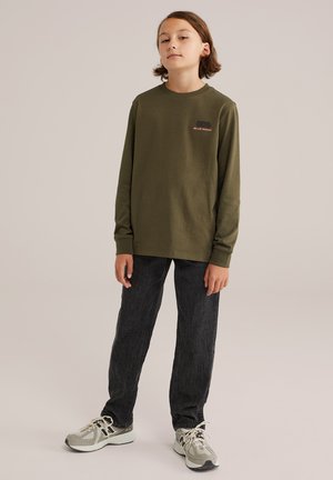 Jeune personne debout, portant une chemise à manches longues olive avec le texte "BLUE RIDGE", un jean noir et des baskets beiges, sur un fond uni.