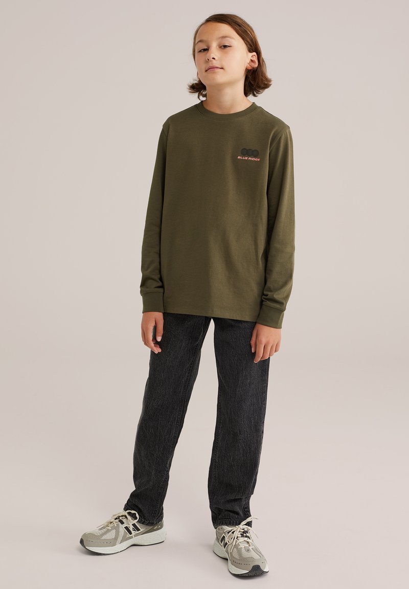 En ung person som står upp, klädd i en olivgrön långärmad skjorta med texten "BLUE RIDGE", svarta jeans och beige sneakers mot en enfärgad bakgrund.