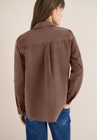 Femme portant une chemise marron à manches longues avec un pli au dos, associée à un jean bleu, debout contre un mur beige.