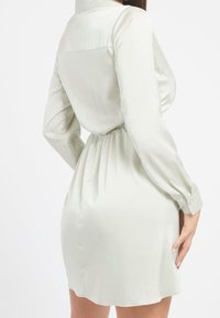 Nainen, joka on pukeutunut pitkähihaiseen, vaaleaan, lyhyeen mekkoon, jossa on kireä vyötärö, seisoo yksi käsi lanteellansa.