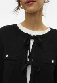 Cardigan noir texturé avec un col rond, fermeture à nœud papillon et deux poches avant avec des accents de boutons argentés. Porté sur un haut blanc.