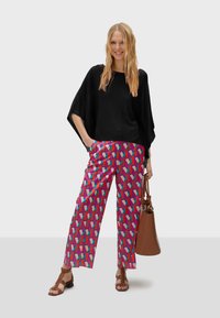 Černý volný top s křídlatými rukávy, doplněný mnohobarevnými kalhotami se vzorem v geometrických tvarech. Hnědá taška a sandály.