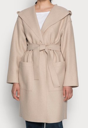 Beige vyöllinen takki, jossa suuri kaulus ja kaksi etutaskua, päällä valkoinen paita ja tummat housut.