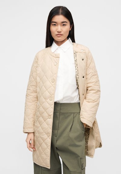 Καπιτονέ μπεζ εξωτερικά ρούχα με κουμπώματα με κουμπιά, με αντίθετη επένδυση με μοτίβο. Φορεμένο πάνω από ένα λευκό πουκάμισο με γιακά και χακί παντελόνια.