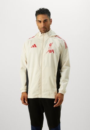 Veste coupe-vent crème clair avec des accents rouges. Logo de Liverpool, fermeture éclair, poignets élastiques et panneaux latéraux noirs.