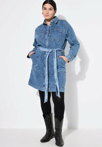 Vestido camisero de denim con cintura ceñida por un cinturón, mangas largas, dos bolsillos en el pecho y terminación con dobladillo desflecado. Combinado con leggings negros y botas de cowboy.