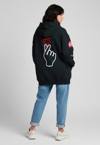 Felpa nera con grande design di una mano bianca e cuore rosso sul retro, con logo rosa sulla manica. Abbinata a jeans arrotolati azzurri chiari e scarpe bianche.