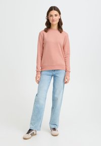 Hele roosa ümmarguse kaelusega dressipluus, soonikkoes mansettide ja äärega, kombineeritud heleroheliste laiade pükstega ja beežide tossudega.