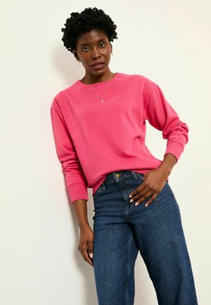 Rosa Sweatshirt aus Baumwolle mit gerippten Bündchen und Halsausschnitt, kombiniert mit hoch taillierten blauen Denim-Jeans. Modell posiert vor einem neutralen Hintergrund.